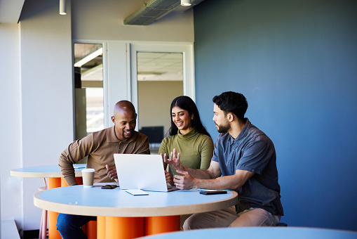 Diverse group of young businesspeople smiling while working together on a laptop at a table in the lounge area of an office