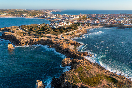 Drone view of rocky peninsula and town of Peniche, Portugal. Summer sunset haze, little foliage and rocky cliffs, peninsula and rocks, fishing town, horizon, ocean waves break on rocky shores.