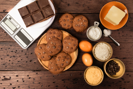 Overhead view of chocolate cookies with the ingredients over rustic wooden table at home in the kitchen. In the picture are the folowing ingredients: chocolate cookies,eggs,chcolate chips,chocolate bars,butter,wheat flour, baking powder, brown sugar and tea spoon with salt.