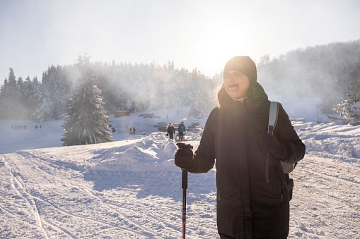 Young cheerful woman in a snowy mountain - ski resort. She is wearing a black jacket, a black knit hat, and black gloves. He holds a pole in one hand.