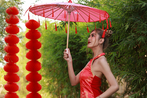 Portrait  beautiful  Asian woman in a Chinese cheongsam qipao dress posing with a beautiful red paper umbrella and red lantern  on a forest background, copy space, Chinese New Year concept