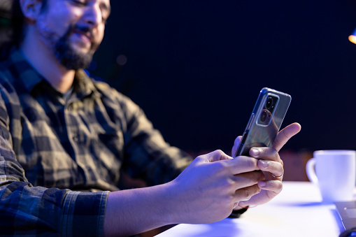 Closeup of young man sitting and using his mobile device for communication, chatting with friends. Male individual grasping his smartphone, browsing the internet, checking social media.