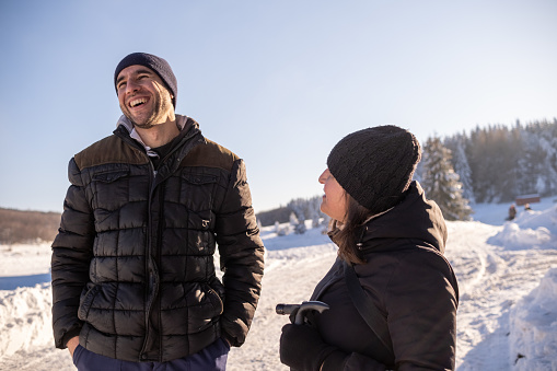 A man and a woman are talking outdoors in a ski resort. The woman looks at the man, and he laughs and looks away.