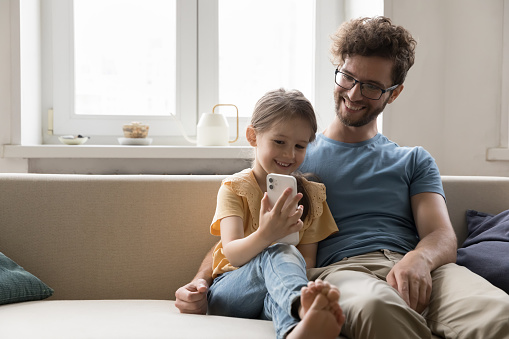 Happy young man sit on cozy couch with little daughter use mobile phone, communicate, play online game. Smiling cute girl enjoy weekend together with father, watch funny video. Modern tech, internet