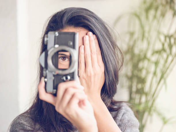 Unrecognizable female with long dark hair looking through retro film photo camera while covering half face with hand and taking photo