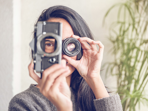 Unrecognizable female with long dark hair looking through hole in retro analog photo camera while covering eye with lens and pretending to take photo