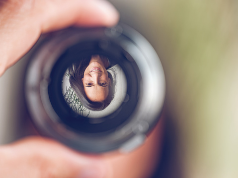 Closeup of crop hand holding lens with upside down reflection of smiling female with long dark hair looking at camera against blurred background