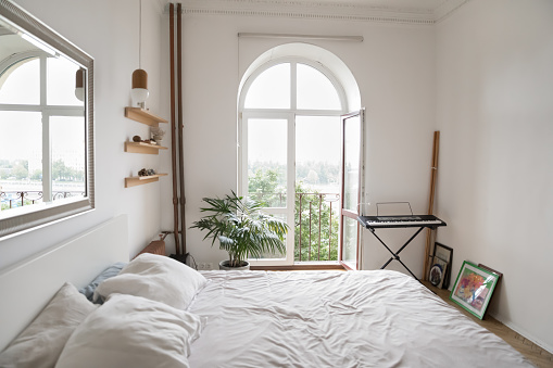 Cozy light bedroom with big mirror on wall, shelves, synthesizer, white wrinkled linen on double bed, view to summer city through opened door, no people. Interior of stylish, cosy modern empty bedroom