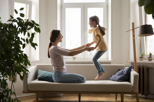 Cheerful mother play with little cute daughter on couch, mom holding hands while joyful girl jumping on soft sofa. Happy family spend time at home, enjoy warm relationship, have fun. Motherhood, love