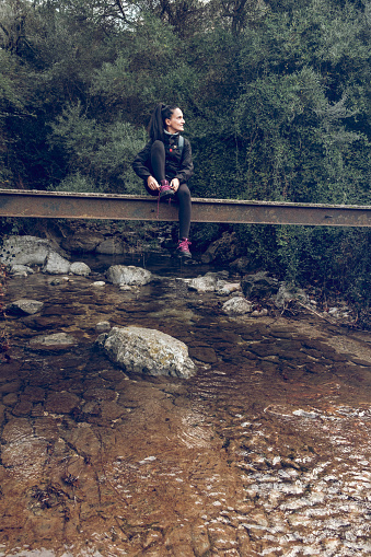 Female tourist in casual clothes sitting on stone bridge above river in forest of Mallorca and looking away