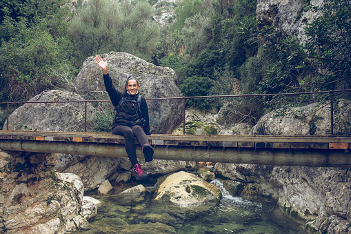 Full length of cheerful young Hispanic female hiker in casual outfit smiling and waving hand while sitting on aged wooden footbridge over river during trekking in Biniaraix ravine