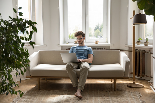 Focused young freelancer man sit on sofa with laptop, work remotely from home office, write email, look through information on internet, spend free time chatting in social media or shopping on-line