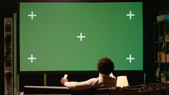 African american man using video projector to binge TV series on subscription based streaming services. Cord cutter enjoying VOD shows on gigantic green screen television set