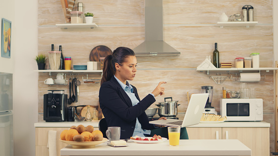 Business woman waving on video call during breakfast. Young freelancer in the kitchen having a healthy meal while talking on a video call with her colleagues from the office, using modern technology and working around the clock
