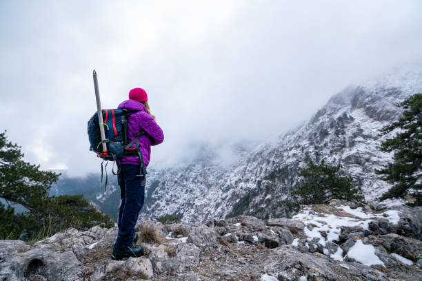 female mountain climber is looking at the landscape and the summit of a high altitude mountain while standing arms crossed. - adventure extreme terrain wilderness area inspiration fotografías e imágenes de stock