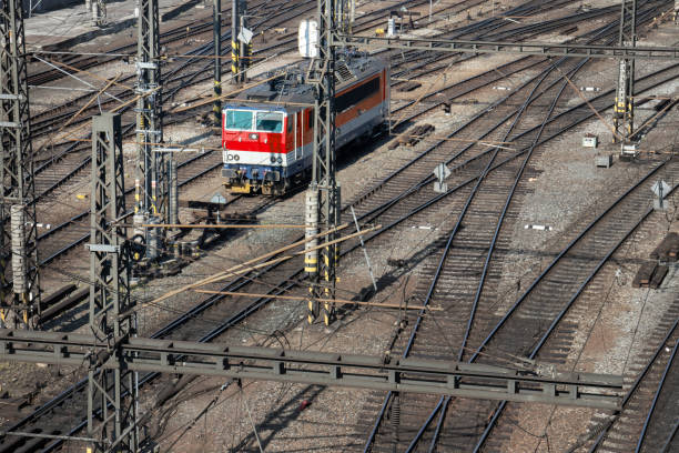 A vibrant train speeds along railway tracks, showcasing a blend of colors while structures support overhead cables. The sunlit surroundings create a bright atmosphere.