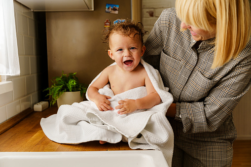 A mother holds her baby wrapped in a towel, the baby has just been bathed in the kitchen sink