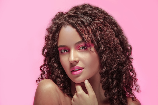 Woman with pink makeup, curly hair, hand on her face, and a smiling expression