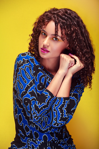 Young woman with an intense gaze and hands on her face, wearing a blue dress, and yellow background