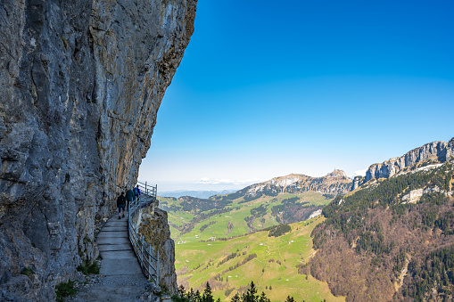 Description: Tourists walk on alpine trail from Äscher to Ebenalp under a big rock wall with Hoher kasten mountin in the background. Seealpsee, Appenzell, Switzerland, Europe.