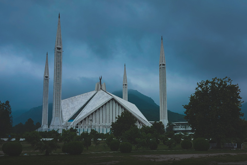 The photo depicts Faisal Mosque in Islamabad, with the mountains in the background. The outdoor scene includes a sky with clouds, trees, and grass.
