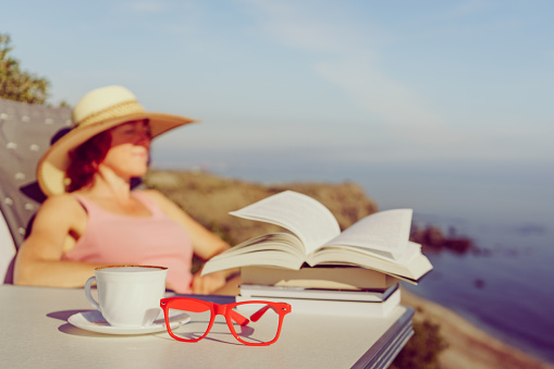 Reading on holidays. Mature tourist woman relaxing on coast, read book, enjoy sea view from cliff. Focus on coffee cup and glasses.