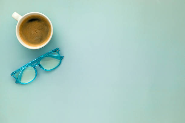 A morning coffee mug and a pair of blue reading glasses on a solid blue background