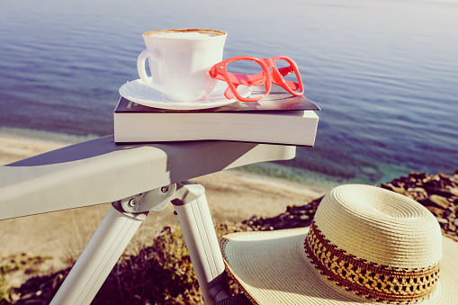 Reading on holidays. Coffee cup and book on chair outdoors against blue sea water background.