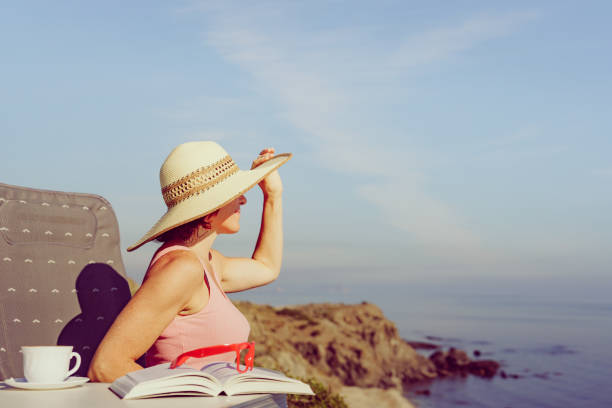 Mature tourist woman relaxing on coast with book and coffee cup, enjoy sea view from cliff. Reading on holidays. Relax time.