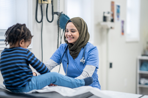 A little boy sits up on an exam table as his Muslim doctor gives him a routine check-up.  The doctor is wearing scrubs and has a hijab on as she checks the young boy over.