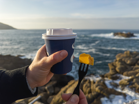 a hand presenting a piece of cheese on a fork in the foreground, with a coffee cup held in the background, all set against the vivid backdrop of the ocean. The blue coffee cup complements the deep blues of the sea, while the yellow cheese adds a pop of color that draws the eye. This scene is not just a snack by the sea; it's a tranquil moment of enjoyment, where the salty air meets the rich aroma of coffee, and the serene ocean waves provide a rhythmic soundtrack to a simple yet perfect experience.