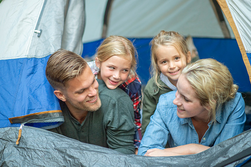 A small family of four are seen laying together in a tent as they. pop their heads out to look around.  They are each dressed casually and smiling as they enjoy the time together.