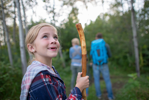 A sweet little girl looks up in to the trees as she spots some birds while on a walk in the woods with her family.  She is dressed in warm layers and holding a walking stick to navigate the terrain.