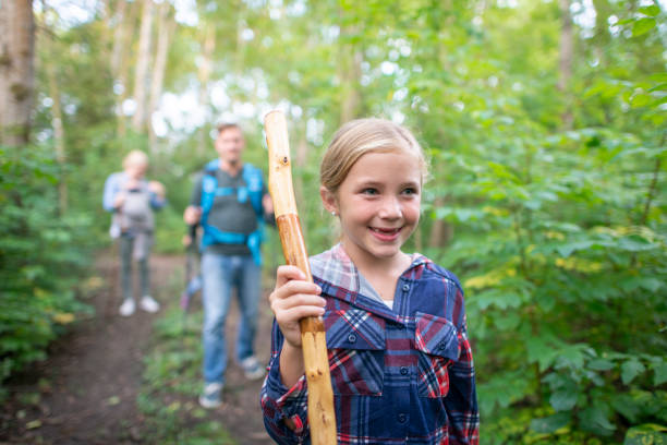 A young girl is seen leading her family on a fall hike.  She is dressed warmly in layers and holding a stick to help her navigate the terrain.