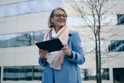 a blond woman with glasses in a coat stands in front of an office building and uses a tablet