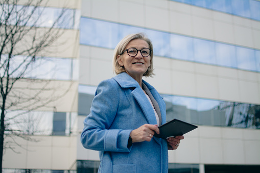 blonde woman with glasses stands in front of office building and using tablet