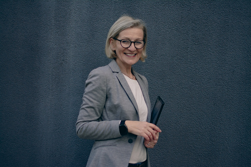 blond mature woman with glasses stands in front of a dark background and uses a tablet