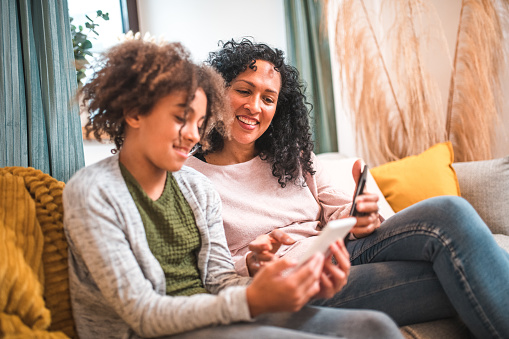 Experience the joy of reading with this endearing image featuring a Hispanic mother and her mixed race daughter. They relax on a sofa in their domestic living room, engrossed in e-books on their e-readers, cherishing the simple pleasure of spending quality time together.