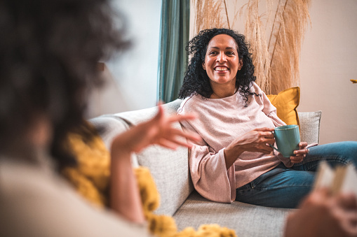 Two friends, mid-adult Hispanic women, sit on a sofa, chat, and share a hot drink in their welcoming home.
