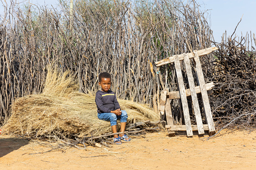 african village child sited on a pile of thatch in front of a branch fence in the yard