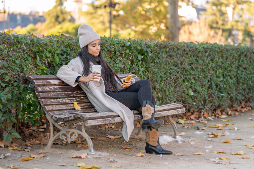 Relaxed woman in a winter outfit sits on a bench in the park, enjoying her phone.