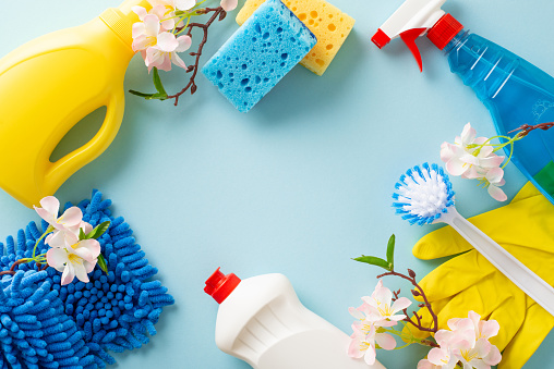 A top view snapshot of cleanliness rituals. Dusters, gloves, flowers and cleaning agents neatly arranged on a soft blue surface. An inviting composition for your promotional text or tagline
