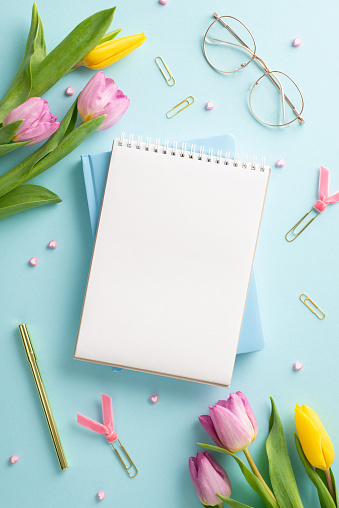Business Blossom: Elevate Women's Day in professional space. Top view of office setup with stylish glasses, essential tools, planners and fresh tulips on a serene pastel blue base with text space