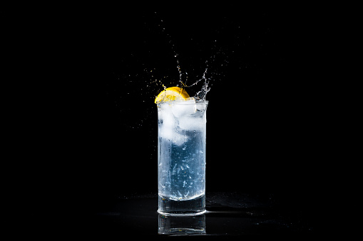 Glass of water and lemon splashing in summer drink. Refreshing drink with splash droplets isolated on black background with copy space.