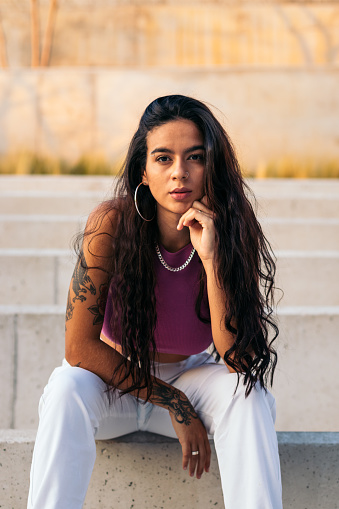 Portrait of a young latina woman with tattoos and piercings sitting on some steps in the city looking at the camera