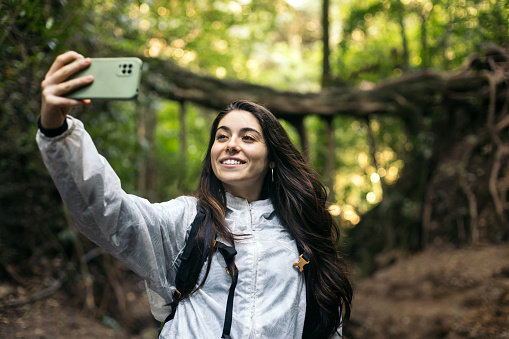 Young woman with raincoat and backpack taking a selfie with smartphone in the jungle during her trip to Costa Rica