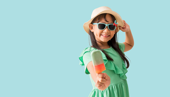 Happy Asian little girl posing with wear a hat with sunglasses holding ice cream, Holiday summer fashion green dress, isolated on pastel blue color background