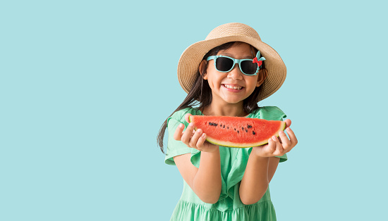 Happy Asian little girl posing with wear a hat with sunglasses holding watermelon slices, Holiday summer fashion green dress, isolated on pastel blue color background