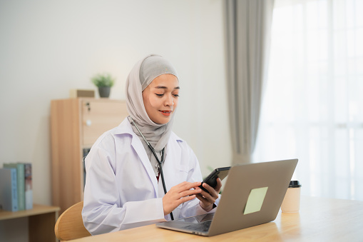 Muslim islam Asian doctor man wearing hijab and stethoscope working with laptop computer and using mobile phone, smartphone. Female doctor work at home office hospital. Health hospital concept.