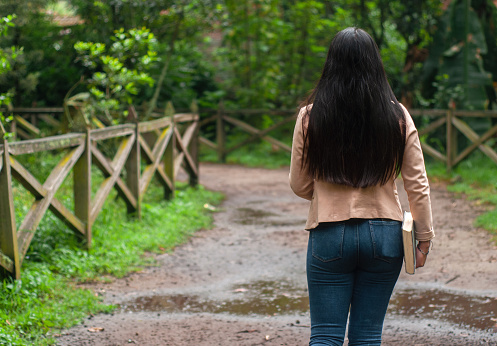 copy space of A Latina woman in blue jeans strolling leisurely along a muddy forest trail with a closed book in her hand, surrounded by greenery and rustic wooden fences.